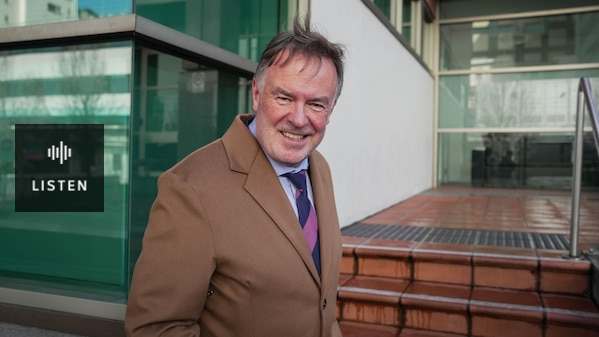 A man in a fawn-coloured coat smiles at the camera. Has Audio.