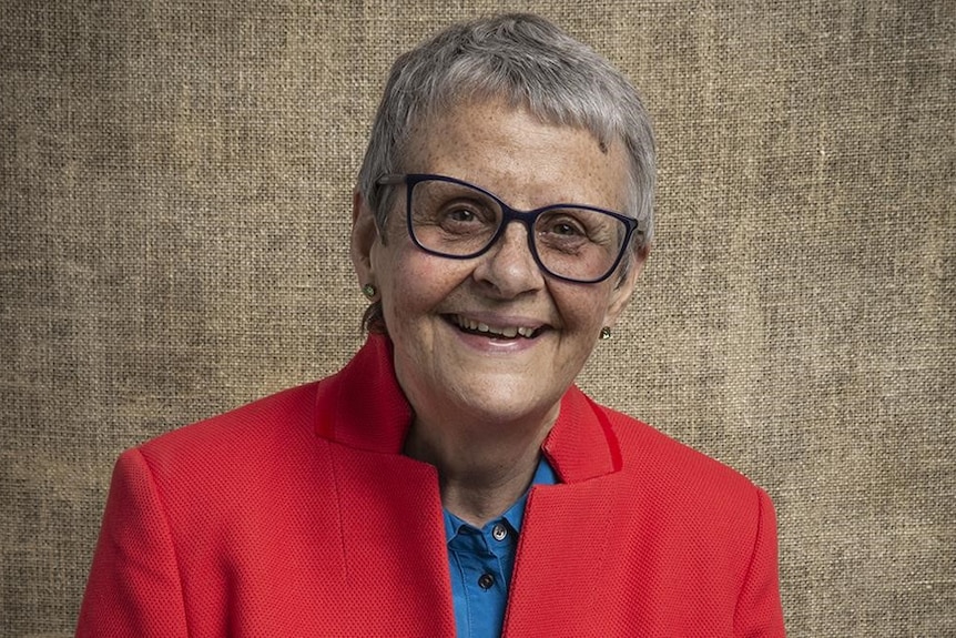 Wendy Mitchell from shoulders up wearing a red jacket and black-rimmed glasses. She has short grey hair and she smiles widely.