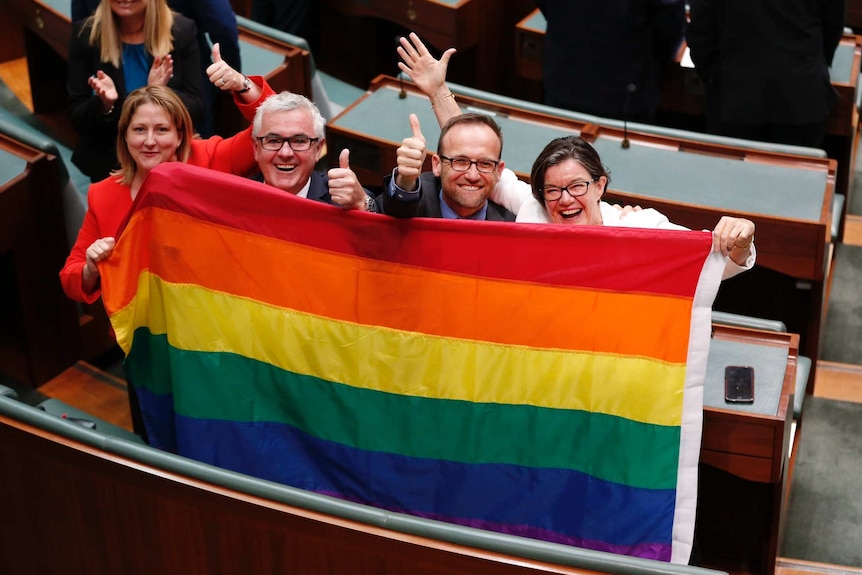 Rebekha Sharkie, Andrew Wilkie, Adam Bandt and Cathy McGowan hold up a rainbow flag. They are smiling and giving the thumbs up