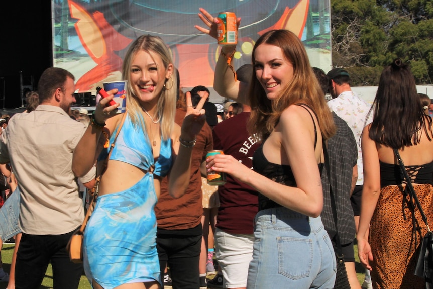 Girls in bright coloured outfits smile at camera in front of live music stage and and crowd. 