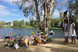 A woman and two childrean stand near a large pile of flowers and a cross on a riverbank.