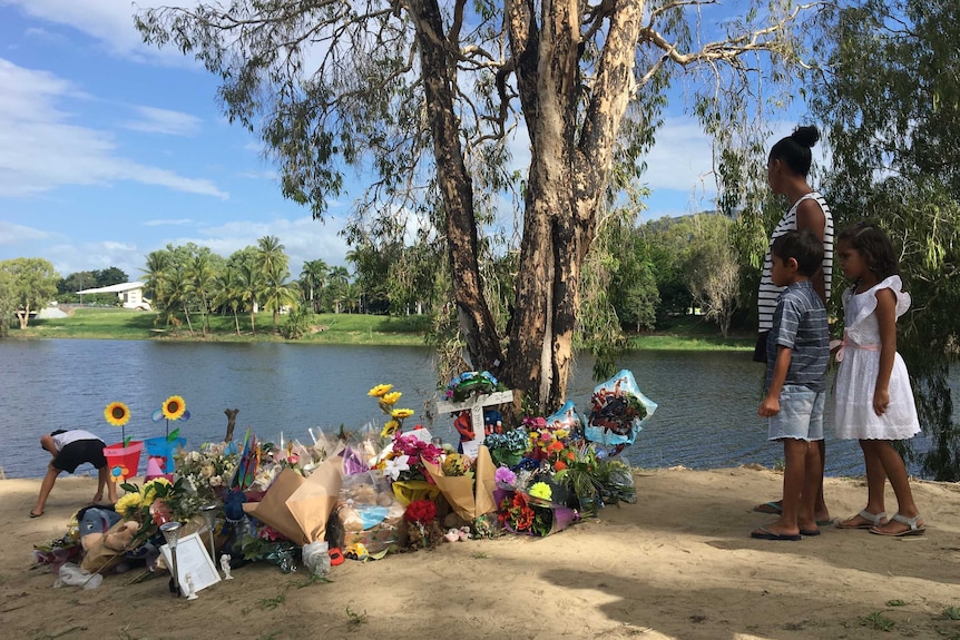 A woman and two childrean stand near a large pile of flowers and a cross on a riverbank.