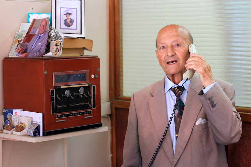 Elderly man speaking on the telephone, standing next to a 1960s telephone exchange machine