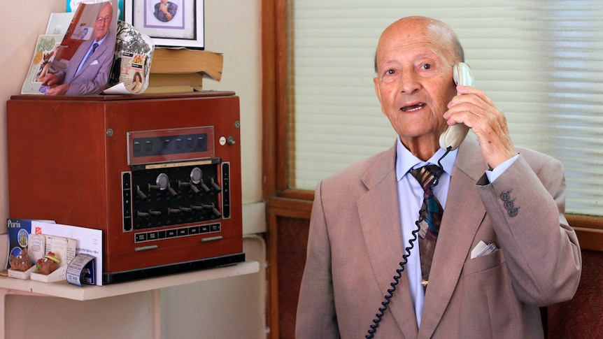 Elderly man speaking on the telephone, standing next to a 1960s telephone exchange machine