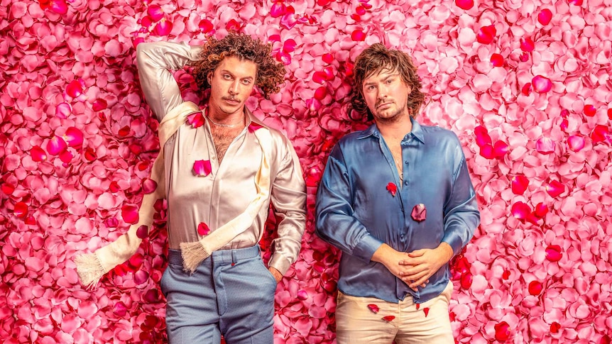 peking duk (two white men) lying on a bed of pink and red rose petals, they're wearing silk shirts (blue and blush colour) and t