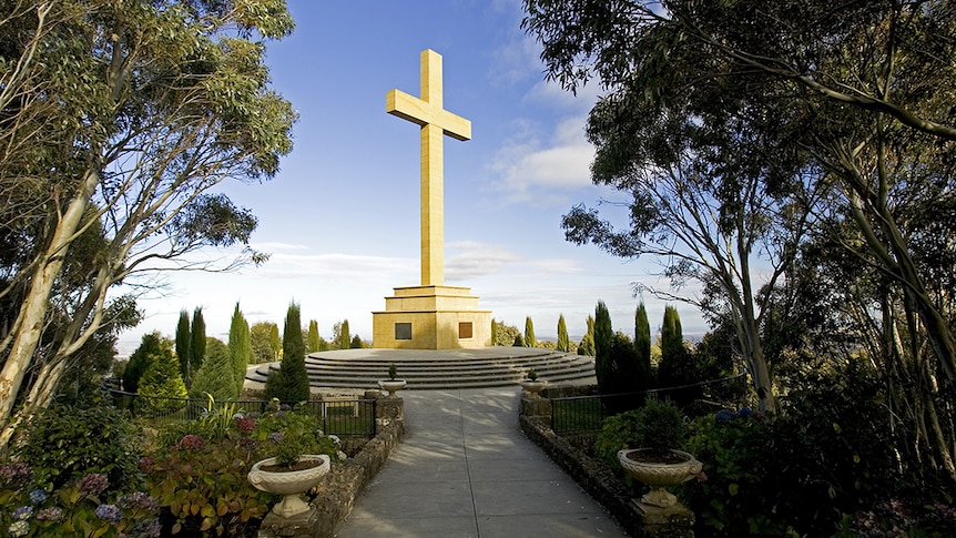 A photo of yellow cross at top of mountain, surrounded by trees 
