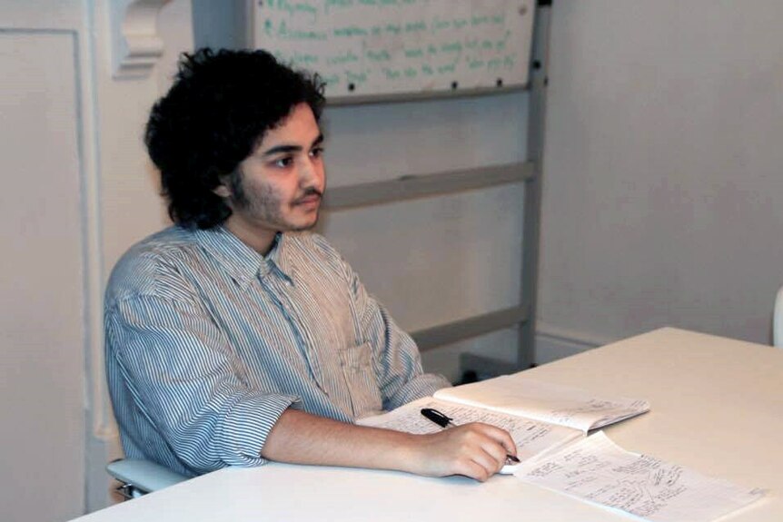 A man in a stripy shirt sits at a desk in a class room, with a writing pad in front of him.