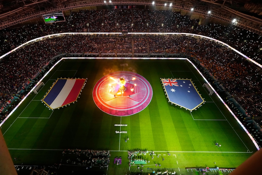 An overhead shot looking down a stadium at night-time, with large French and Australian flags on the pitch and a light show.