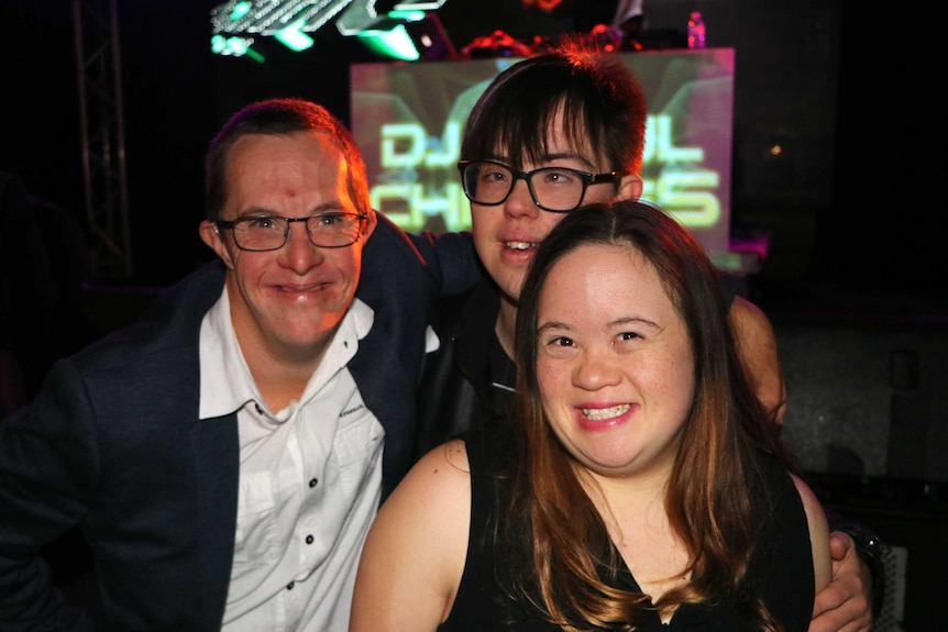 Two men and a woman smile on the dance floor, bright lights behind them.