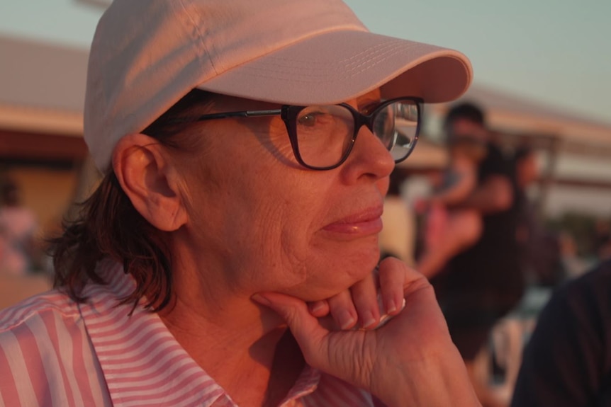 A sunset close-up of a woman wearing glasses and a cap, resting her chin on one hand, looking pensive.