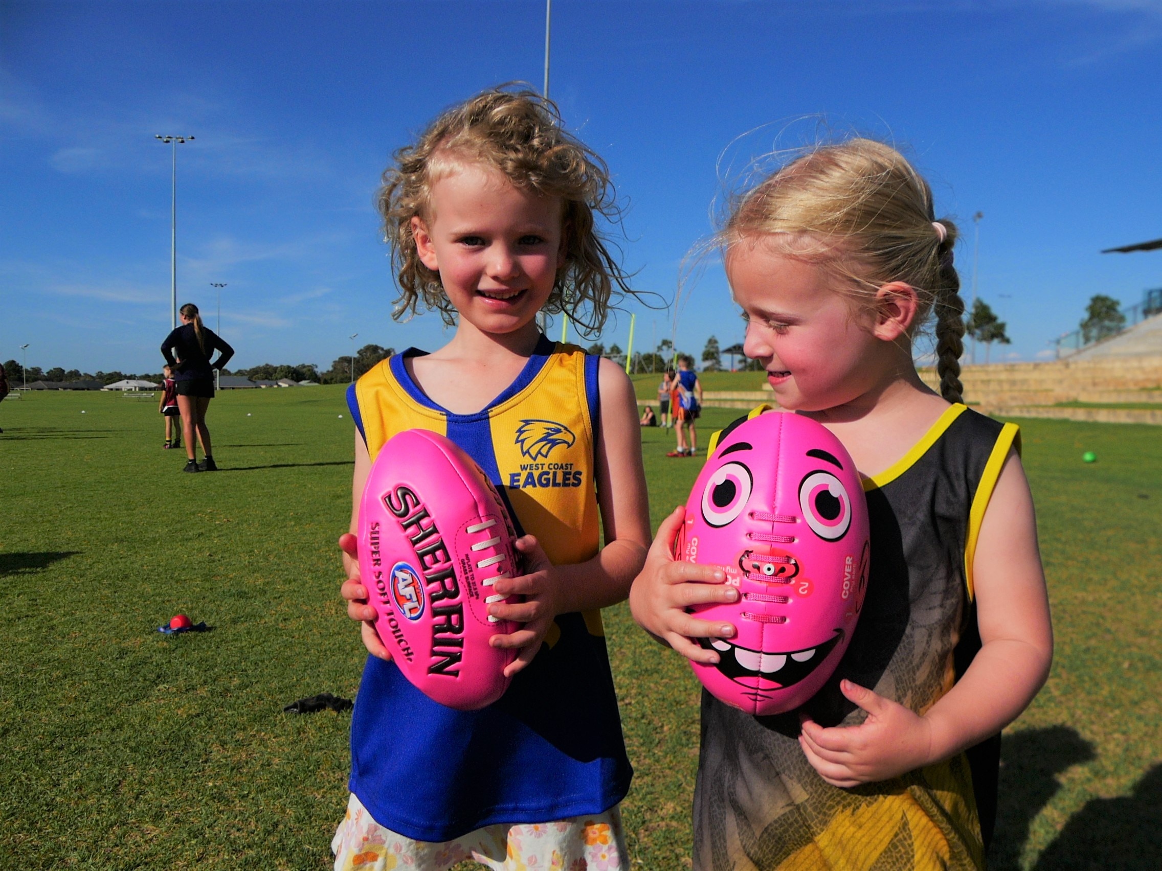 Two small children with blonde hair stand next to each other holding pink footballs. 