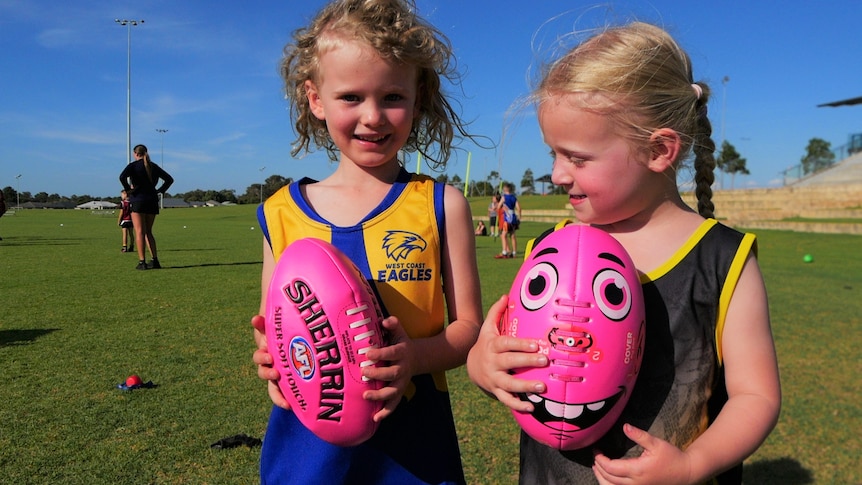 Two small children with blonde hair stand next to each other holding pink footballs.