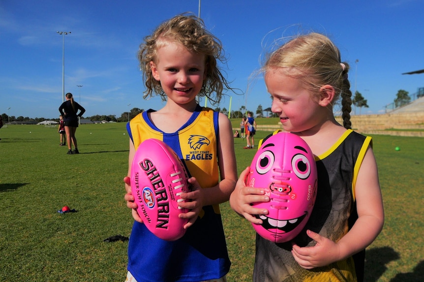 Two small children with blonde hair stand next to each other holding pink footballs. 