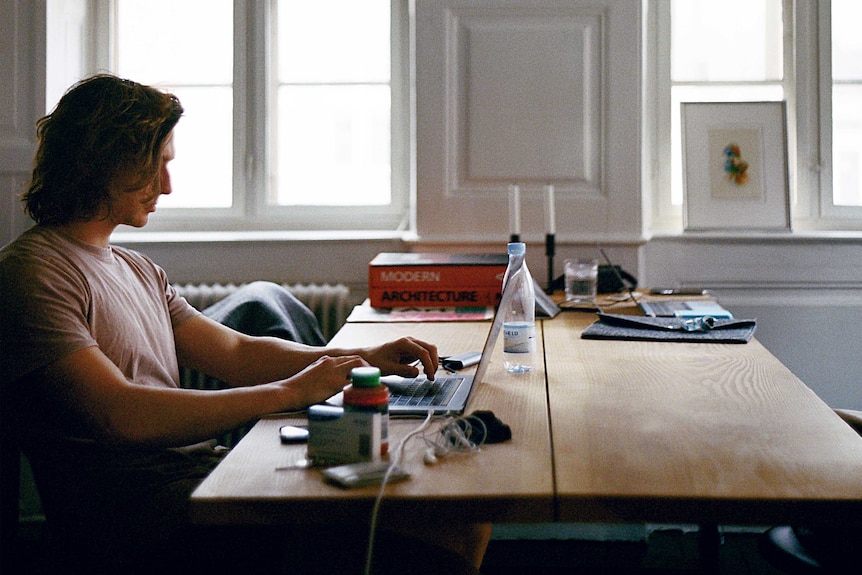 A man sits at a slightly cluttered table at home typing on a laptop.