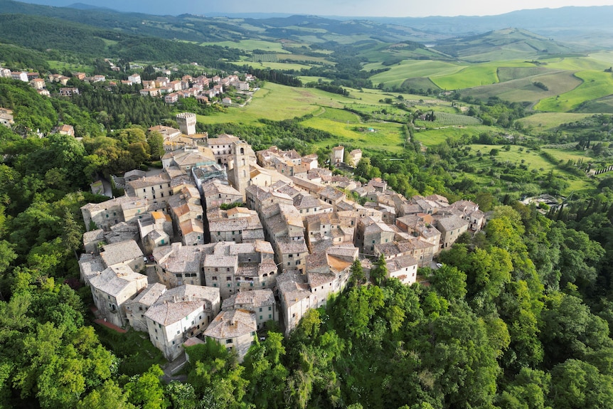 An aerial view of an ancient Tuscan village nestled amongst bright green trees on a country hillside.