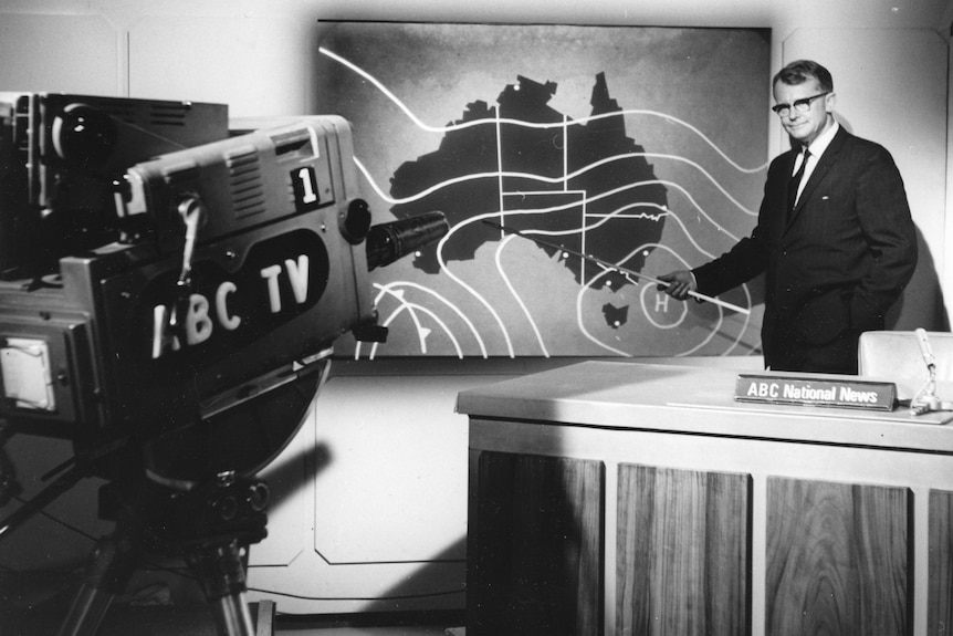 A black and white photo of a 1960s weather presenter pointing to a map of Australia, with