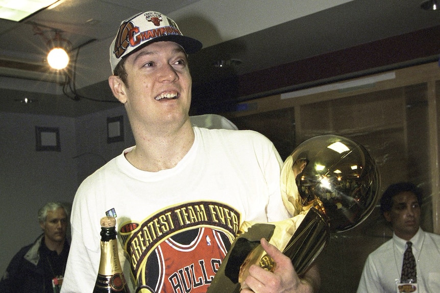 Luc Longley wears a tshirt and cap holding the 1996 NBA championship trophy and a bottle of champagne
