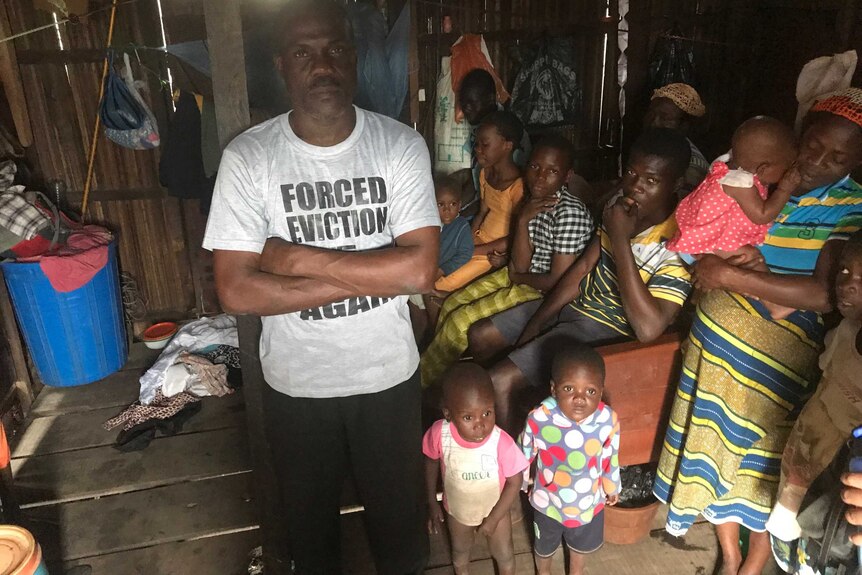 A man in a t-shirt that says forced eviction has crossed arms in a crowded shanty living room