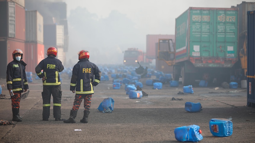 Firefighters look on  as smoke and fumes fill the air and plastic containers are spread over the ground.