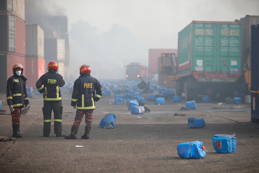 Firefighters look on  as smoke and fumes fill the air and plastic containers are spread over the ground.