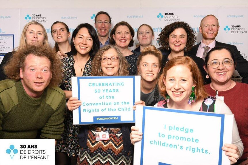 A group of donor-conceived people huddle together in a group holding up signs and posing for a photo smiling.