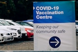 A sign reads 'COVID-19 Vaccination Centre: Keeping our community safe' in front of parked cars.