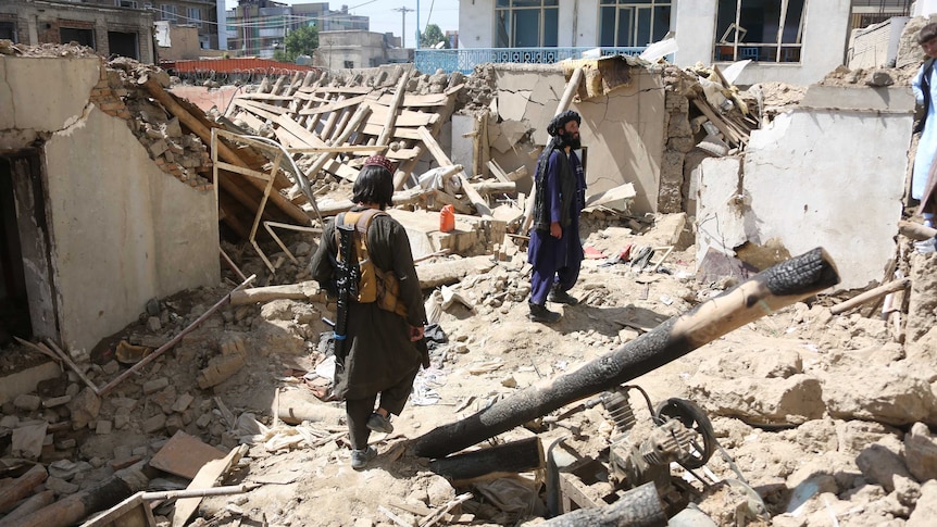 two men, one with a gun on his back, stand in a destroyed building, among pipes and rubble