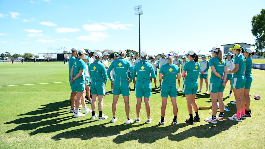 Australia's women's cricket squad, all wearing warm up gear, stand in a circle on the field on a bright sunny day