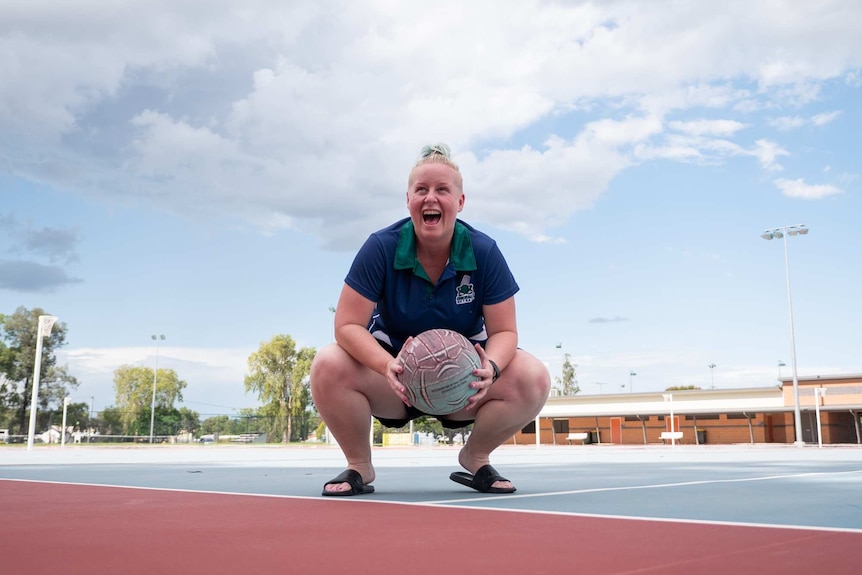 Kristy Serotzki crouches down on the netball courts holding a netball between her knees while laughing