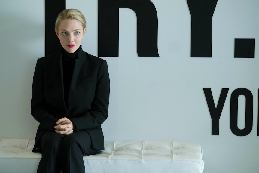 A young woman with blonde hair and red lipstick dressed in a black turtleneck and suit sits on a white couch in an office.