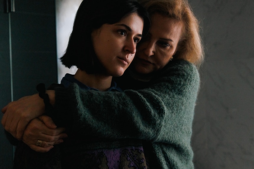 A woman with blond hair wraps her arms around her daughter, in a portrait taken in a dark shadowy room.