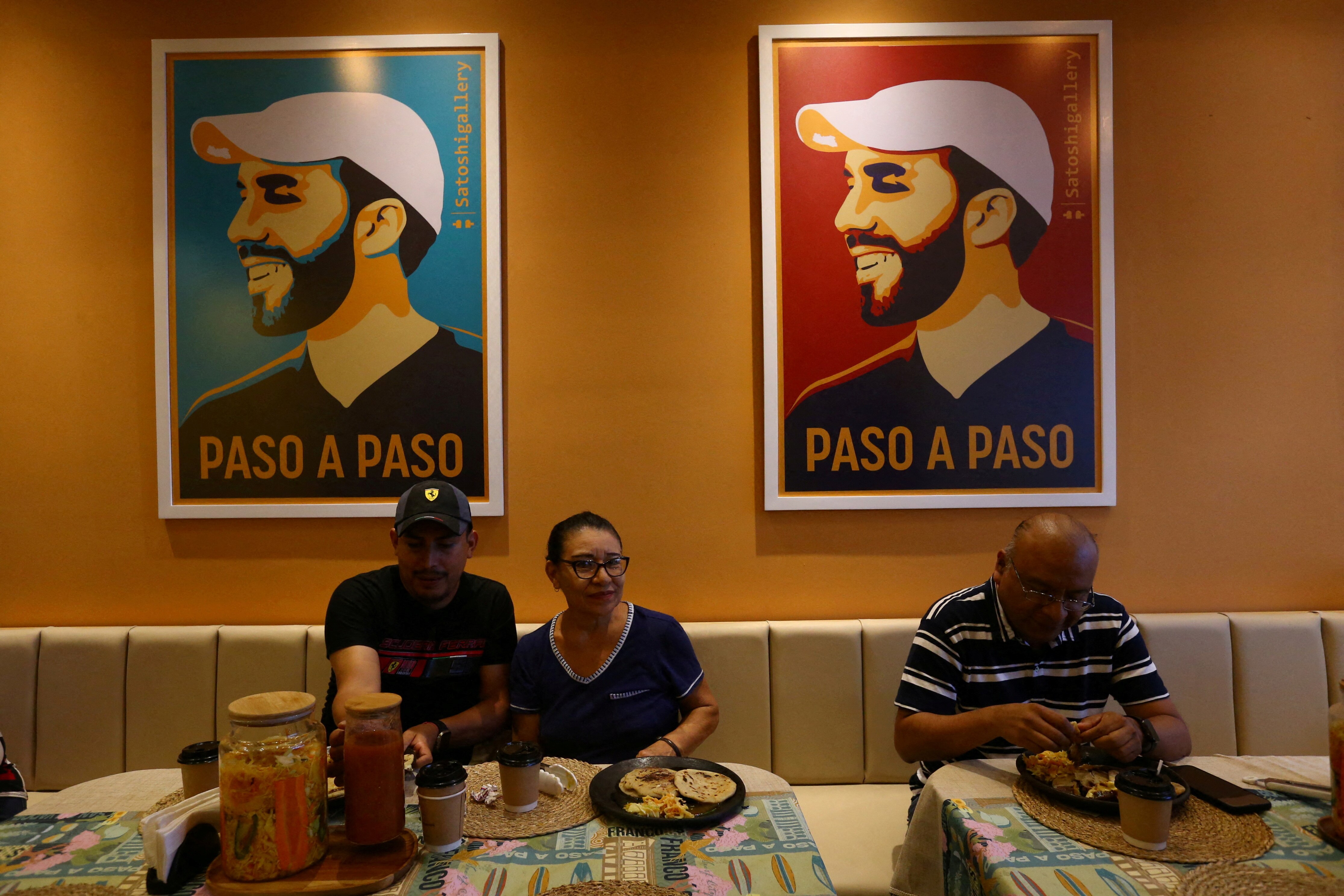 La gente se sienta bajo dos pinturas de colores brillantes de Nayib Bukele en un restaurante.