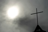 A cross sits on top of a church in Berlin with the sky and sun in the background.