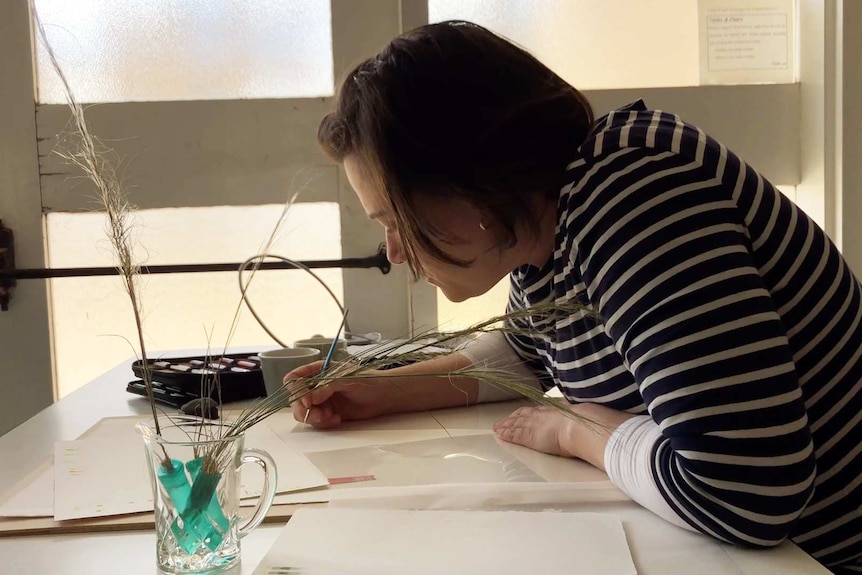 A woman hold a paintbrush, leaning over a piece of paper.