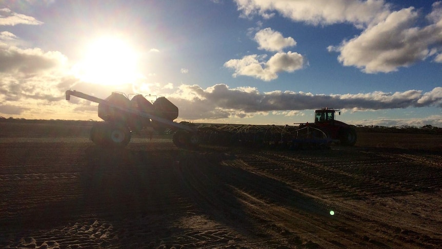 A tractor pulls an airseeder and bar in a dry paddock in Western Australia as the sun sets behind the machinery