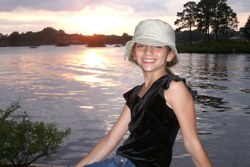 A teenage girl in a bucket hat smiles at the camera in front of a lake