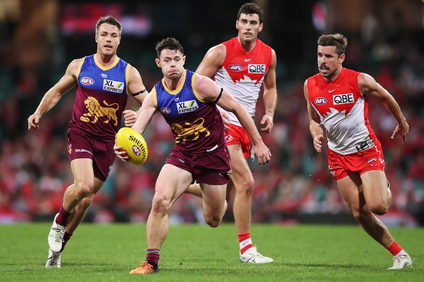 A Brisbane AFL player holds the ball in one hand as he runs, trying to get clear of a Sydney defender.