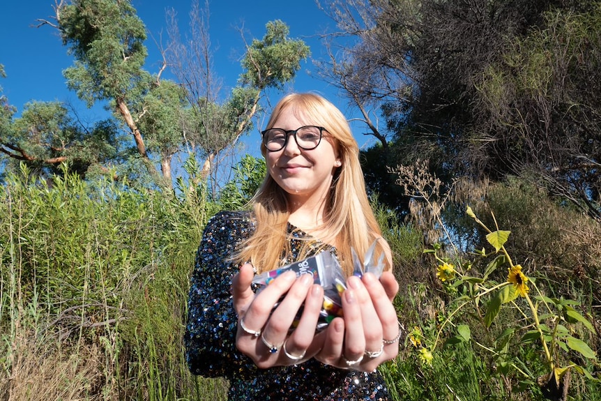 A young woman with long blonde hair holds forward her hands full of packets of rainbow beads.