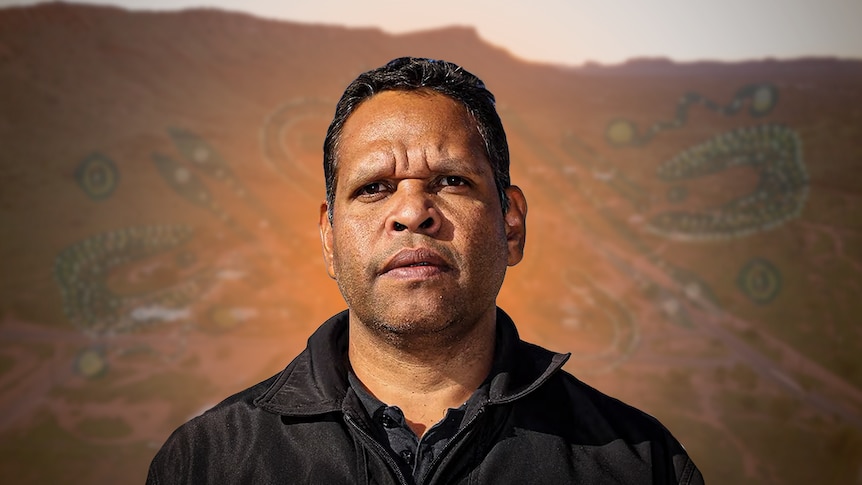 A man wearing a black shirt looks seriously off-camera. Behind him is faint Aboriginal print and the town of Alice Springs.