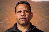 A man wearing a black shirt looks seriously off-camera. Behind him is faint Aboriginal print and the town of Alice Springs.