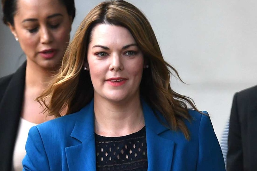 A woman in a blue jacket walks, flanked by people in suits.