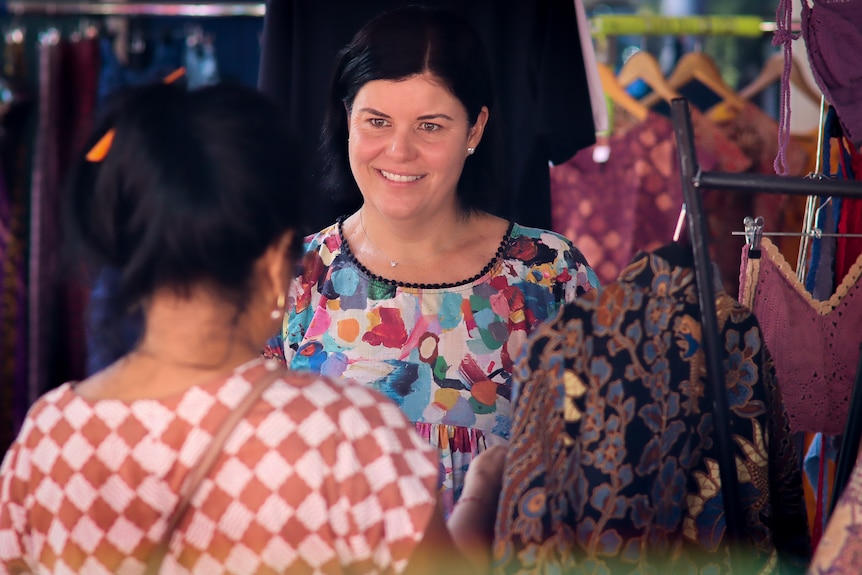A woman with short dark hair smiles as she talks to a woman at a market stall.