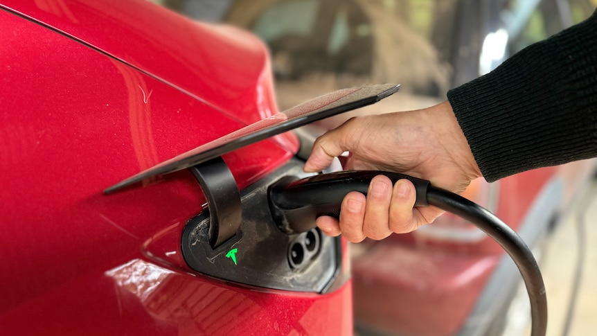 A hand holds an electric vehicle charging plug against a car.