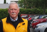 A man wearing a yellow vest stands in a car park full of cars