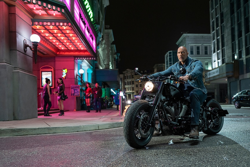 A man sits on motorcycle on the street at night time outside neon lit cinema.
