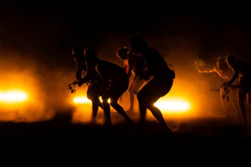 People dance by firelight with tree leaves.