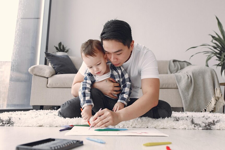 A father sits on the floor with his young son and draws on a piece of paper.