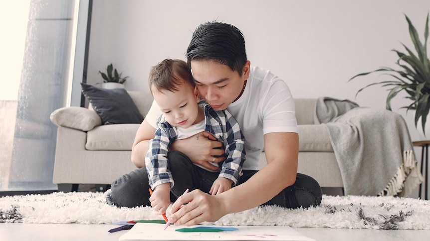 A father sits on the floor with his young son and draws on a piece of paper.