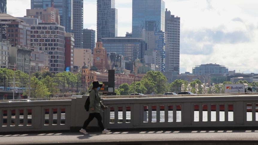 A woman wearing a jacket, backpack and mask crosses a Melbourne bridge.