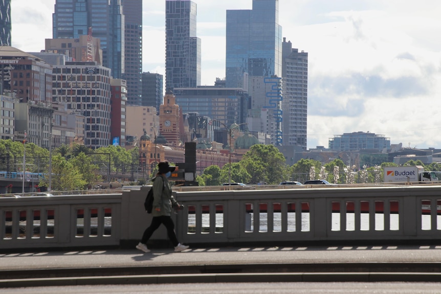 A woman wearing a jacket, backpack and mask crosses a Melbourne bridge.